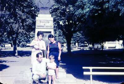 Retrato de plano entero de dos mujeres con Vestido una de ellas con un Bebé en brazos, un hombre con Traje y un niño, todos posando de frente junto a estatua de Teodoro Schmidt en Plaza de Armas de Temuco