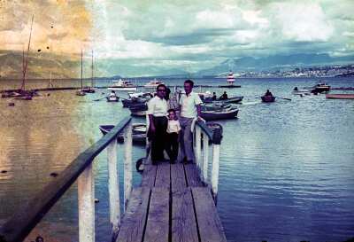Retrato de plano entero de dos hombres y un niño de pie posando de frente en muelle con botes y lanchas ancladas a los lados