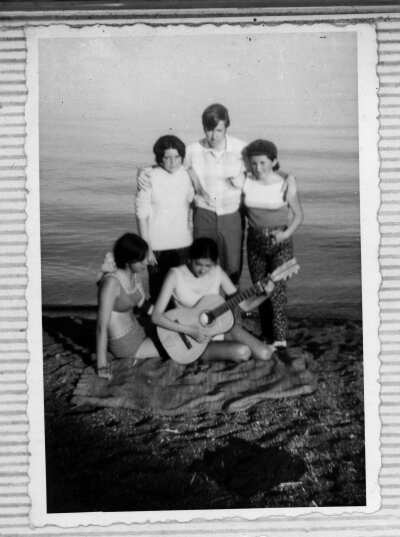 Grupo de personas sentadas y de pie en la orilla del lago, sobre una manta en la piedrecilla. Una de las mujeres sentadas toca la guitarra.
