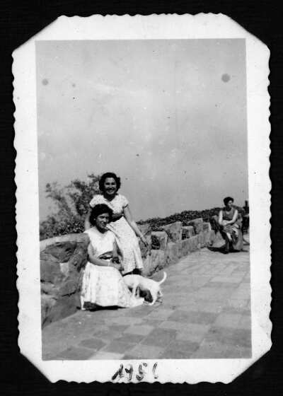 Retrato grupal de dos mujeres sentadas en una barrera de piedra. Ambas visten vestidos y están con un perro. Al fondo, hay otra mujer sentada,