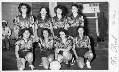 Retrato grupal de plano entero del club deportivo bancario de baloncesto, formadas sobre la cancha. En sus poleras llevan bordadas las iniciales del club. En el fondo, se encuentra el marcador del partido. Se encuentran en el interior de un recinto no identificado