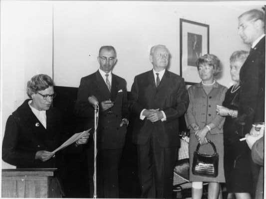 Plano entero de Olga Boettcher leyendo un documento junto a un micrófono, mientras un grupo de Hombres y Mujeres las acompañan. En la pared del fondo, se encuentra colgado un retrato del presidente de la época, señor Pedro Aguirre Cerda. Se encuentran en el interior de una infraestructura no identificada