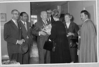 plano general de una mujer saludando a un hombre de la mano. Lleva consigo una cartera y un ramo de flores. A su lado los acompaña un grupo de Hombres y Olga Boettcher. Se encuentran en la inauguración del nuevo edificio del colegio Alemán de La Unión