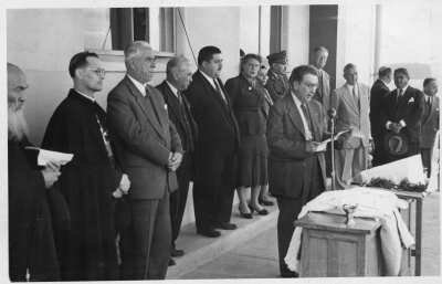 plano general de un hombre leyendo un documento frente a un micrófono. A sus espaldas, formados en una hilera, se encuentra dos sacerdotes, tres hombres, Olga Boettcher, una mujer, un carabinero y otro grupo de hombres. Todos le acompañan en la inauguración del hospital Dr. Juan Morey de La Unión realizado en el frontis del edificio