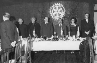 plano general de un grupo de personas, formadas en una hilera detrás de una mesa. La primera mujer de izquierda a derecha es Olga Boettcher. Se encuentran en el interior de la sede del Rotary club de La Unión durante un banquete