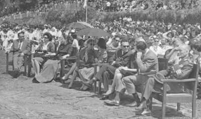 plano general de Olga Boettcher, primera gobernadora de La Unión y Eva Sámano, primera dama de México, conversando sentadas bajo la sombra de un quitasol. A su alrededor, hay un grupo numeroso de personas sentadas en el interior del estadio Carlos Vogel, mientras se lleva a cabo un homenaje a la primera dama durante su visita a la ciudad de La Unión