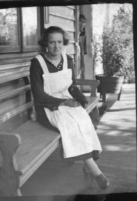 Retrato de plano entero de una mujer con Vestido, posando sentada en una banca. En el fondo, hay otra mujer asomada desde la puerta de la casa.