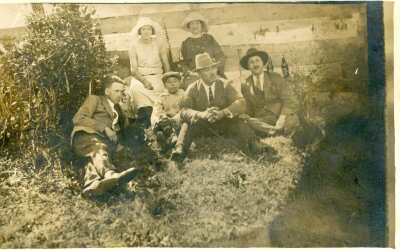 Retrato grupal de plano entero de Hombres, Mujeres y un niño posando sentados sobre el pasto. A sus espaldas hay un cerco de madera