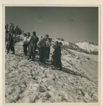 plano general de un grupo de Hombres y Mujeres reunidos frente a unas tablas de esquí las cuales están enterradas en la nieve. La segunda mujer de izquierda a derecha es Olga Boettcher. Se encuentran de paseo en unas montañas nevadas no identificadas.