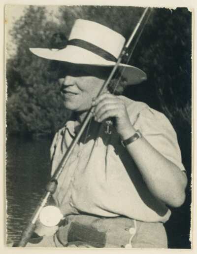 Plano medio largo de Olga Boettcher sosteniendo una caña de pescar. A sus espaldas hay árboles y un río no identificado.