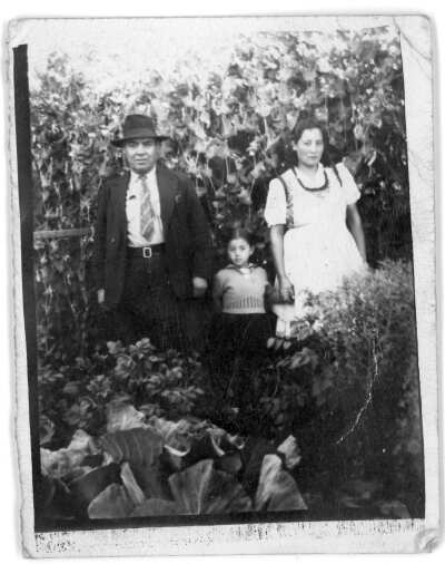 Retrato de plano entero de un hombre mayor y una mujer, posando mientras toman la mano de una Niña. Se encuentran parados entre unas plantas, bajo las ramas de unos árboles.