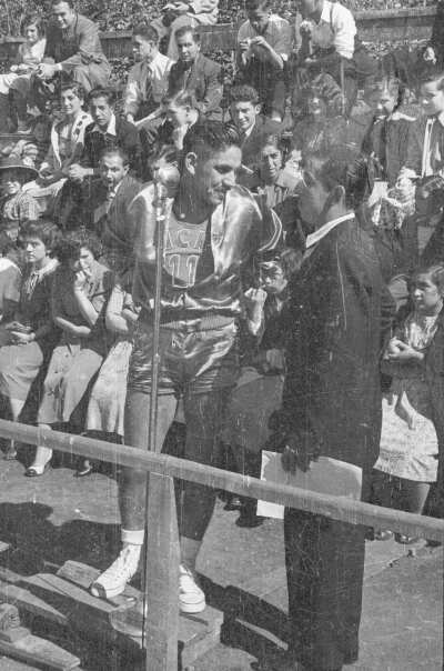 Plano entero de dos hombres conversando, uno está vestido de traje y sostiene un papel en su mano, el otro es jugador de baloncesto, detrás de ellos, hombres y mujeres sentados en las graderías se encuentran observando el partido.