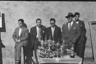 Plano entero de un grupo de cinco hombres apoyados en una pared detrás de la mesa que tiene los trofeos del juego