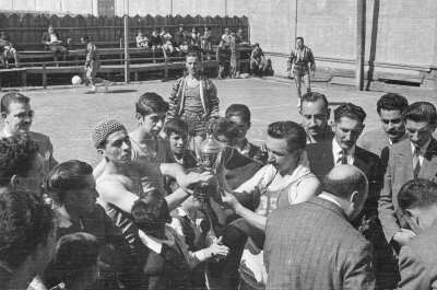 Plano entero de cinco hombres sosteniendo un trofeo, entre ellos hay un niño observándolos, a su alrededor hay un grupo de seis hombres reunidos conversando, mientras que están rodeados de dos mujeres y cuatro hombres que observan el trofeo. En el fondo se observan personas en las graderías mientras que un hombre persigue un balón.