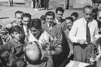 Plano entero de jugador de equipo masculino conversando con un grupo de niños con el trofeo en sus manos, en su cuello lleva puesta una toalla. Alrededor lo observan un grupo de hombres vestidos con traje.