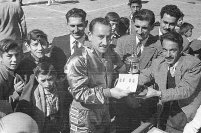 Retrato grupal de cinco hombres y tres niños, uno de los hombres le entrega al jugador de baloncesto un trofeo y un diploma, detrás de ellos hay cinco niños.