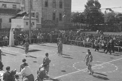Plano general de seis hombres jugando un partido de baloncesto, a su alrededor se ven las graderías repletas de personas observando.