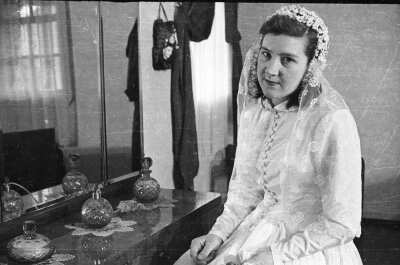 Retrato de plano medio de una mujer vestida de novia posando sentada junto a un mueble tocador en el dormitorio de una casa