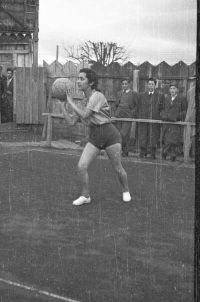 -Plano entero de una mujer jugadora de baloncesto parada sobre la cancha de juego con una pelota en la mano a prontas de dar un pase. A su alrededor hay un grupo de Hombre 