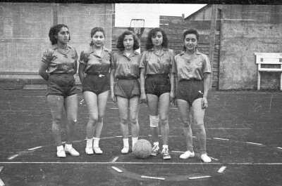 Retrato grupal de plano entero de mujeres jóvenes, todas ellas son jugadores de un equipo de baloncesto, en el medio se puede apreciar a una de ellas con una rodilla vendada. Todas están posando de pie con la pelota en el piso en el interior de una cancha
