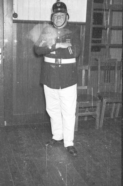 Retrato de plano entero de 1HM posando de pie con los brazos cruzados mientras usa su uniforme de bombero. Está en el interior de una infraestructura.