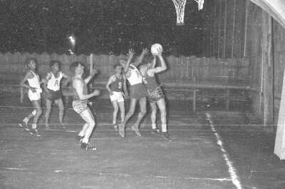 plano general de dos equipos de baloncesto masculino disputando la pelota bajo el aro. Ambos equipos llevan en sus poleras escrito números o la letra A'. Se encuentran jugando en el interior de una cancha durante la noche.