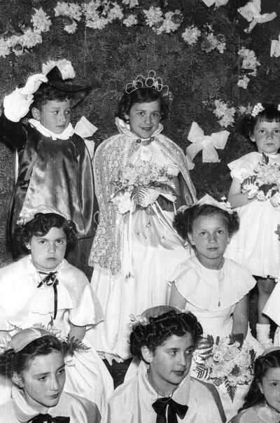 Plano entero de un niña coronada a reina sentada con un arreglo floral de gran tamaño a sus espaldas. Junto a ella hay un niño disfrazado usando un sombrero y un grupo de Niñas sentadas con un ramo de flores en sus manos. Están en el interior de una escuela durante una jornada de actividades artísticas.