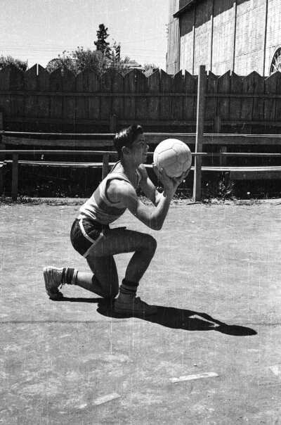 Plano entero de un jugador con una rodilla en suelo, realizando un tiro con en balón en sus manos. Es perteneciente a un club deportivo de baloncesto masculino, no identificado. Está en el interior de una cancha.