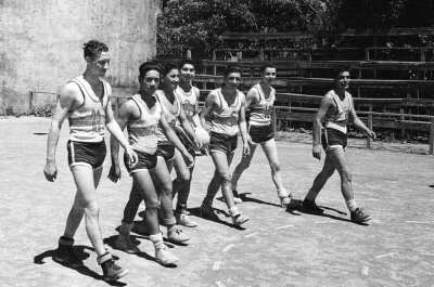 Plano entero de un grupo de jugadores caminando juntos. Son integrantes de un club deportivo de baloncesto masculino, no identificado. Están en el interior de una cancha.