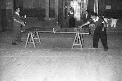 plano general de dos hombres jugando tenis de mesa. En el fondo hay dos hombres observándolos durante el partido en el interior de un local