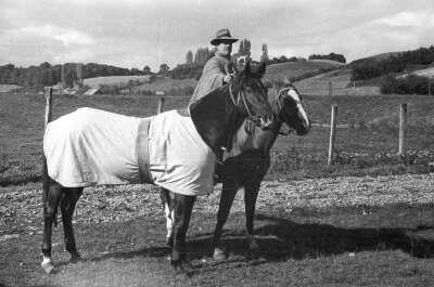 Plano entero de un hombre montado a caballo mientras le agarra las riendas a un caballo que está junto a ellos. está a un costado de un camino de ripio en el campo
