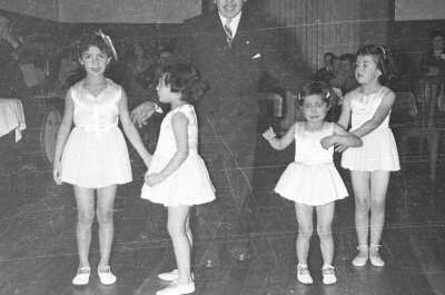 Plano entero de cuatro niñas caracterizadas como bailarinas de ballet paradas junto a un hombre vestido de Traje. En el fondo hay un grupo de Hombres y Mujer sentados alrededor de unas mesas en el interior de un local