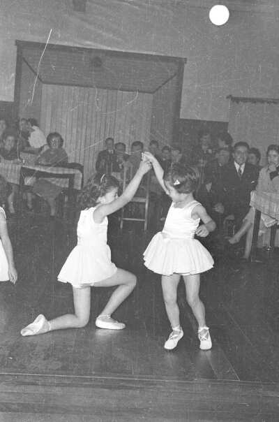 Plano entero de dos niñas caracterizadas como bailarinas de ballet, bailando tomadas de la mano. A su alrededor hay un grupo de Hombres y Mujer que las observan sentados alrededor de unas mesas en el interior de un local
