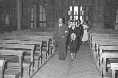 Plano entero de una mujer vestida de novia caminando tomada del brazo de un hombre mayor. Tras ellos lo siguen un grupo pequeño de mujeres y hombres, mientras avanzan por la nave central de la Parroquia San José, rumbo al altar.