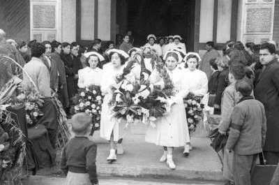 Plano entero de un grupo de mujeres vestidas de blanco, saliendo de la parroquia con una corona de flores en sus manos. A su alrededor hay un grupo de Niños, Mujer y Hombres que están esperando la salida del féretro en las afueras de la Parroquia San José