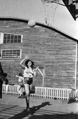 Plano entero de una jugadora de baloncesto de un equipo no identificado, lanzando un balón hacia un aro. En un costado, hay un hombre vestido de Traje que la está observando. Están en el interior de una cancha