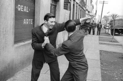 Plano medio largo de dos hombres jugando a las luchas sobre la vereda. En el fondo se acercan tres hombres vestido de Traje y un niño con una bicicleta. Los hombres se encuentran en el frontis de un estudio fotográfico