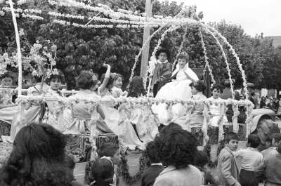Plano entero de la pareja de reyes de la escuela superior de Niñas Nº2, mirando hacia la cámara. Van acompañados de un grupos de niñas en el interior de un carro alegórico, el cual está decorado por flores y guirnaldas. Caminan junto a ellos un grupo de Niños y Niñas por la calle pavimentada en el frontis de la plaza de La Concordia