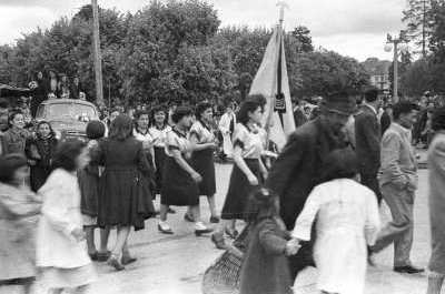 plano general de una niña levantando una bandera por el mástil, mientras le siguen un grupo de Niñas vestidas todas de igual manera. Tras ellas, le siguen un grupo de mujeres paradas en la parte trasera de una camioneta mientras avanzan junto a otro grupo de Mujer, Hombres y Niños, desfilando por un costado de la Plaza de La Concordia