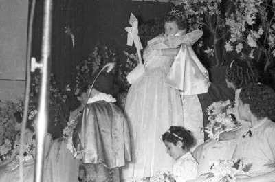 Plano entero de una mujer con vestido abrazando a una Niña vestida de reina. A su alrededor un Niño y un grupo de Niñas las observan. Se encuentran en el gimnasio de la escuela de Niñas Nº2 durante una jornada de actividades escolares