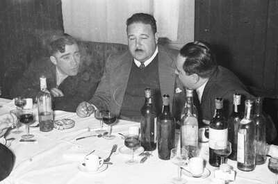 Plano medio de tres hombres de Traje conversando sentados junto una mesa. El del medio tiene un cigarro encendido en su mano mientras la afirma sobre la mesa la cual tiene botellas de licores encima. Se encuentran en el interior de un local durante una celebración