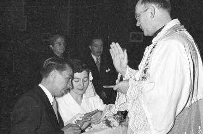 Plano entero de un sacerdote realizando una señal con su mano, frente a una pareja de novios, quienes se encuentran en un reclinatorio, hincados frente a él. En el fondo una mujer mayor y un hombre los acompañan, durante la celebración de su matrimonio religioso, llevado a cabo en la parroquia San José