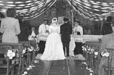 Plano entero de una pareja de novios, parados en el altar, de espaldas a la cámara. Frente a ellos, Se encuentra un sacerdote y tres acólitos. En cada costado, se encuentran sus invitados. Están en el interior de una parroquia no identificada, durante una misa de celebración de matrimonio