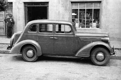 -Plano entero de un auto estacionado a un costado de la vereda, en el frontis del estudio fotográfico --foto Real-- donde un hombre con sombrero se encuentra apoyado en la puerta continua de la entrada de la infraestructura-