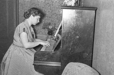 Plano medio largo de Frederica von Edelsberg sentada tocando un piano de pared. Frente a ella, se encuentra abierta una partitura. En el fondo, hay un florero junto a la pared. Está en el interior de una casa.