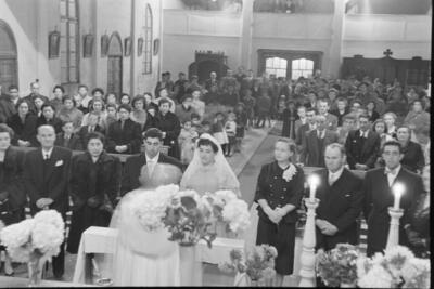 plano general de un grupo numeroso de Mujeres, Hombres, Niños y Niñas, parados junto a sus bancas en dirección al altar, donde se encuentra un sacerdote parado frente a una pareja de novios y sus padrinos, realizando la misa de celebración de su matrimonio en el interior de la parroquia San José
