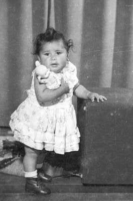 Retrato de estudio de Plano entero de una niña con Vestido, de temprana edad, posando junto a un asiento con una muñeca en sus brazos