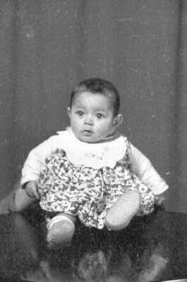 Retrato de estudio de Plano entero de una Bebé, con Vestido y babero, posando sentada mientras una persona escondida la sostiene por la espalda