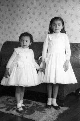 Retrato de plano entero de dos niñas con Vestido posando tomadas de las manos junto a un sillón. La Niña de la izquierda está festejando su cumpleañosen el interior de una casa