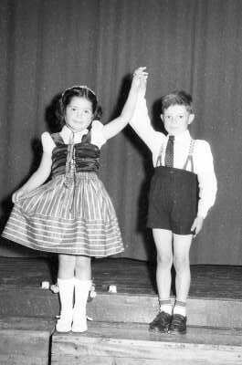 Retrato de plano entero de una niña y un niño posando tomados de la mano en los peldaños de una tarima ubicada en el interior del gimnasio de la escuela superior de niñas Nº2